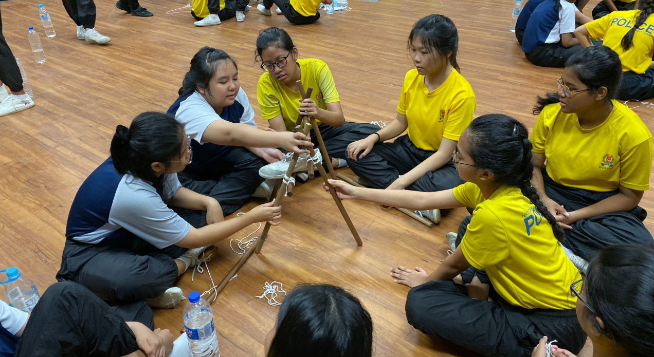 Students in yellow shirts sit on a wooden floor, working together to build a structure using wooden sticks and rope.